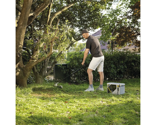 Mann bei der Gartenpflege mit einem elektrischen Rasentrimmer und einer tragbaren Powerstation auf einer Rasenfläche unter Bäumen.