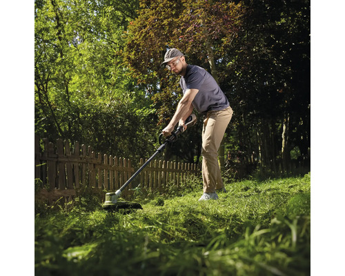 Ein Mann arbeitet mit einem Rasentrimmer an einem Holzzaun in einem grünen, sonnigen Garten.