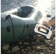 Eine Hand hält eine tragbare elektrische Luftpumpe mit Lithell Logo beim Aufpumpen eines Schlauchbootes an einem steinigen Ufer am Wasser.