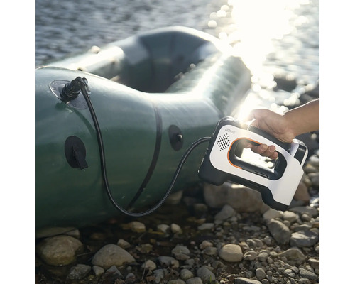 Eine Hand hält eine tragbare elektrische Luftpumpe mit Lithell Logo beim Aufpumpen eines Schlauchbootes an einem steinigen Ufer am Wasser.