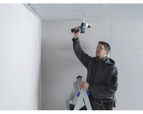 Ein Handwerker montiert eine Überwachungskamera mit einem Akkuschrauber an eine Decke, während er auf einer Leiter steht.
