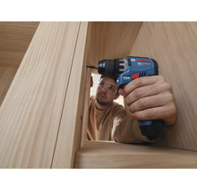Eine Person mit Schutzbrille dreht mit einem Bosch Akku-Bohrschrauber eine Schraube in eine Holzkonstruktion aus hellem Holz.