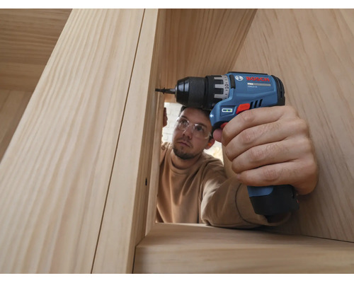Eine Person mit Schutzbrille dreht mit einem Bosch Akku-Bohrschrauber eine Schraube in eine Holzkonstruktion aus hellem Holz.