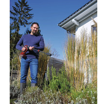 Ein Mann nutzt einen Akku-Druckreiniger im Garten neben hohen Ziergräsern und einem Wohnhaus.