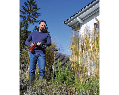 Ein Mann nutzt einen Akku-Druckreiniger im Garten neben hohen Ziergräsern und einem Wohnhaus.