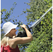 Eine Frau schneidet eine dichte Hecke mit einer Akku-Heckenschere in einem hellen, sonnigen Garten.
