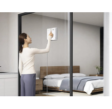 Eine Frau nutzt einen automatischen Fensterputzroboter an einer großen Glastrennwand in einem modern eingerichteten Wohn- oder Schlafzimmer.