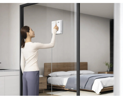 Eine Frau nutzt einen automatischen Fensterputzroboter an einer großen Glastrennwand in einem modern eingerichteten Wohn- oder Schlafzimmer.