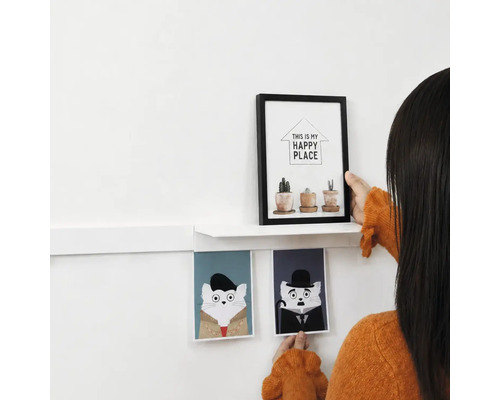 Eine Person dekoriert eine Wand mit einem weißen Wandregal, einem Bilderrahmen mit Pflanzenmotiv und zwei Katzenbildern als Wanddekoration.