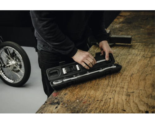 Eine Person öffnet einen schwarzen Kunststoffkoffer mit einem Drehmomentschlüssel und Steckschlüsseleinsätzen auf einer Werkbank aus Holz.