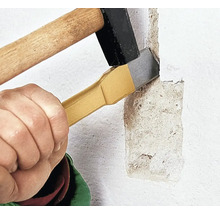 Eine Hand hält einen Flachmeißel an eine verputzte Wand, während ein Hammer auf das Ende des Meißels schlägt, um den Putz abzutragen.