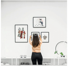Frau hängt Bilderrahmen mit Tiermotiven an eine weiße Wand über einem Sideboard in einer modernen, hellen Wohnzimmerumgebung.
