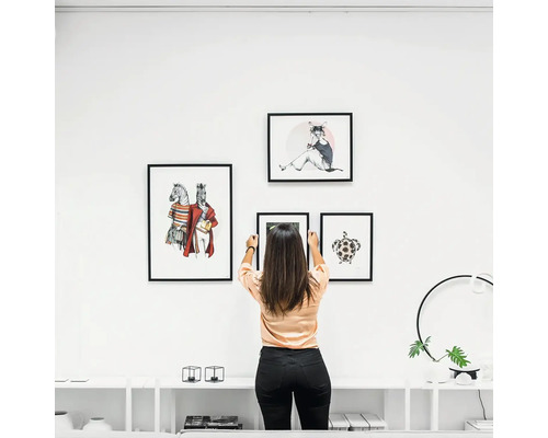 Frau hängt Bilderrahmen mit Tiermotiven an eine weiße Wand über einem Sideboard in einer modernen, hellen Wohnzimmerumgebung.