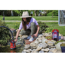 Frau bedient eine Einhell Akku-Klarwasserpumpe an einem hellen Gartenteich mit Steinen und Pflanzen.