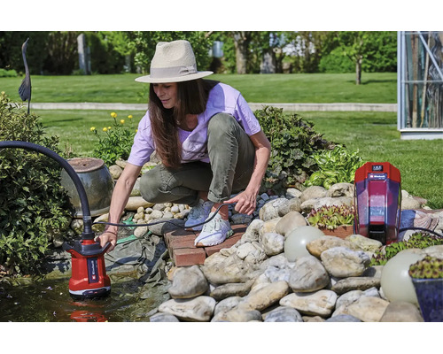 Frau bedient eine Einhell Akku-Klarwasserpumpe an einem hellen Gartenteich mit Steinen und Pflanzen.