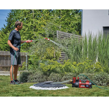 Ein Mann bewässert Pflanzen im Garten mit einer roten Einhell Akku-Gartenpumpe, die Wasser aus einer Zisterne fördert.