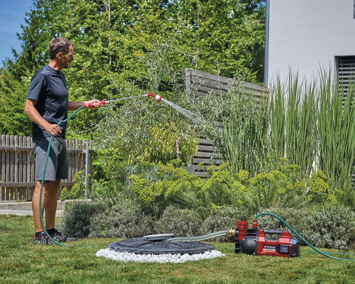 Ein Mann bewässert Pflanzen im Garten mit einer roten Einhell Akku-Gartenpumpe, die Wasser aus einer Zisterne fördert.