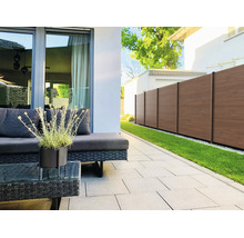 Moderne Terrasse mit dunklen Rattan-Gartenmöbeln, hellen Pflastersteinen und einem braunen Sichtschutzzaun neben einer Rasenfläche.