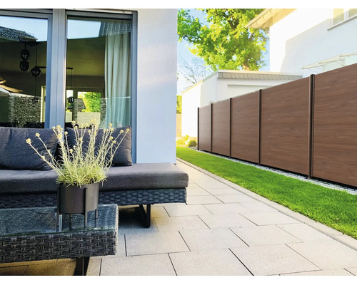 Moderne Terrasse mit dunklen Rattan-Gartenmöbeln, hellen Pflastersteinen und einem braunen Sichtschutzzaun neben einer Rasenfläche.