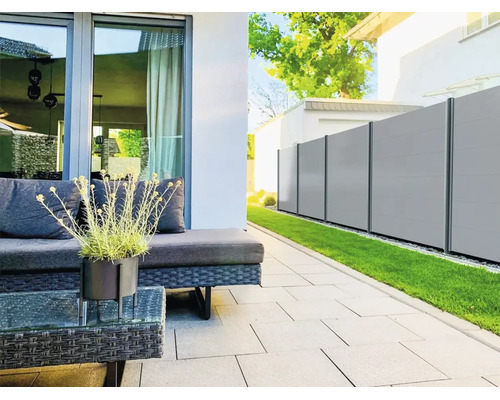 Moderner Sichtschutzzaun aus hellgrauen Paneelen mit dunklen Pfosten in einem Garten neben einer Terrasse mit Loungemöbeln und Rasen.
