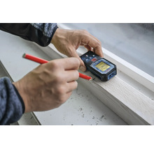 Person verwendet ein digitales Laserdistanzmessgerät und einen Bleistift zur präzisen Markierung an einer Holzleiste auf einer Fensterbank.