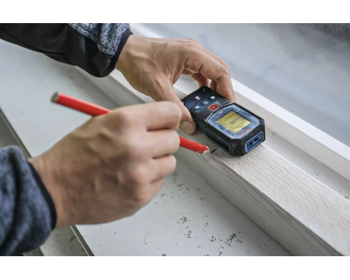 Person verwendet ein digitales Laserdistanzmessgerät und einen Bleistift zur präzisen Markierung an einer Holzleiste auf einer Fensterbank.