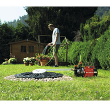 Einhell Gartenpumpe angeschlossen an Zisterne, im Hintergrund bewässert ein Mann Pflanzen in einem hellen Garten.