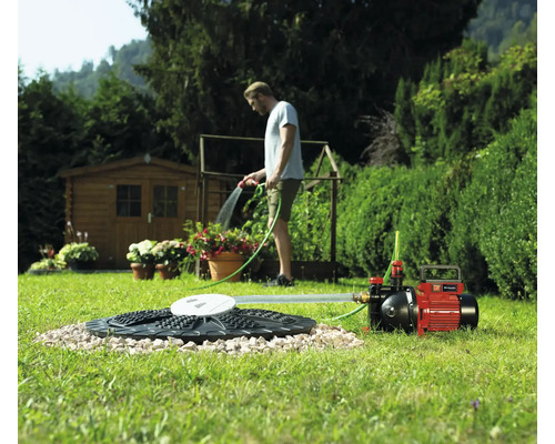 Einhell Gartenpumpe angeschlossen an Zisterne, im Hintergrund bewässert ein Mann Pflanzen in einem hellen Garten.