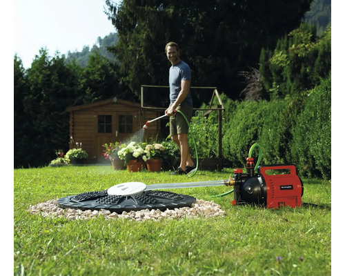 Ein Mann gießt Pflanzen mit einer Einhell Gartenpumpe, die an eine Zisterne im Rasen angeschlossen ist. Ein Gartenhaus steht im Hintergrund.