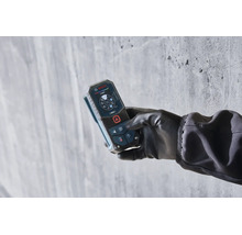 Eine Hand mit Handschuh hält ein Bosch Laser-Entfernungsmessgerät mit digitalem Display vor einer Betonwand.