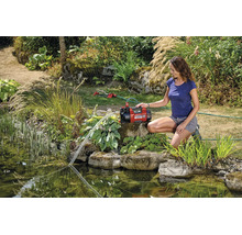 Eine Frau bedient eine rote Einhell Gartenpumpe am Gartenteich zur Bewässerung des Gartens mit einem Rasensprenger im Hintergrund.