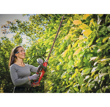 Eine Frau schneidet mit einer Akku-Stab-Heckenschere eine hohe, grüne Hecke im Garten. Sie trägt Schutzbrille und Handschuhe.