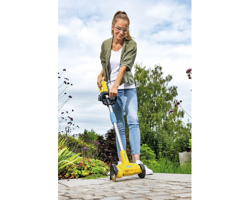Eine Frau reinigt mit einer elektrischen Fugenbürste die Pflasterfugen auf einer Terrasse im Garten.