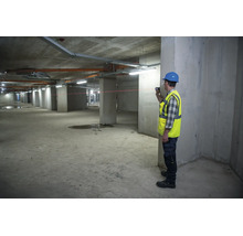 Ein Bauarbeiter mit Schutzhelm und Warnweste verwendet einen Laser-Entfernungsmesser auf einer Betonbaustelle.