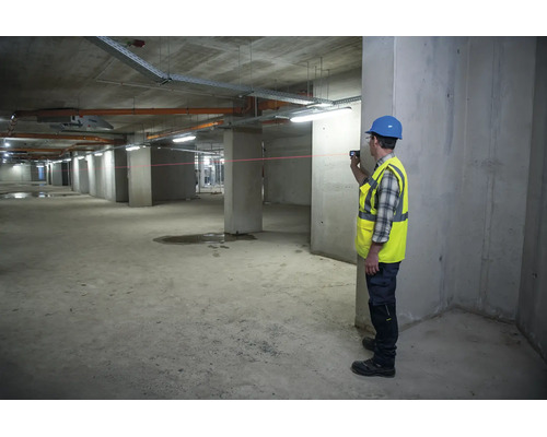 Ein Bauarbeiter mit Schutzhelm und Warnweste verwendet einen Laser-Entfernungsmesser auf einer Betonbaustelle.