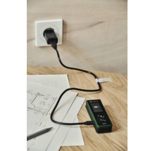 Ein Bosch Zamo Laserentfernungsmesser wird mittels USB Kabel an einer Steckdose auf einem Holztisch mit Bauplan geladen.