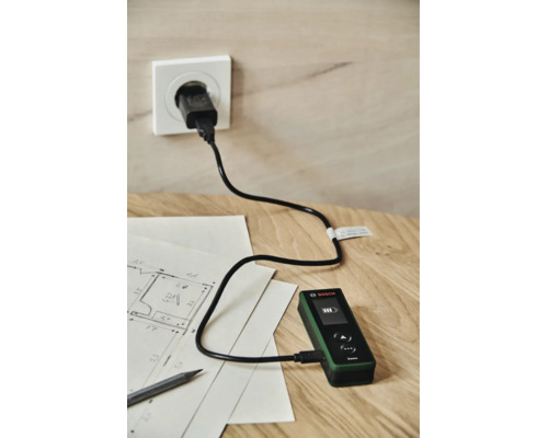 Ein Bosch Zamo Laserentfernungsmesser wird mittels USB Kabel an einer Steckdose auf einem Holztisch mit Bauplan geladen.