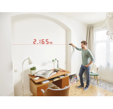 Ein Mann nutzt einen Laser Entfernungsmesser zur Wandvermessung in einem hellen Arbeitszimmer.