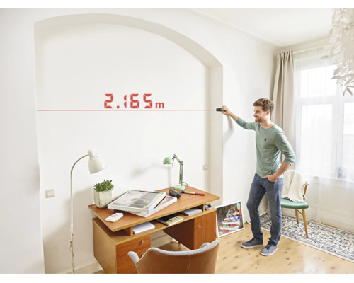 Ein Mann nutzt einen Laser Entfernungsmesser zur Wandvermessung in einem hellen Arbeitszimmer.