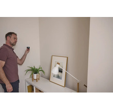 Ein Mann verwendet ein Ortungsgerät an einer Wohnzimmerwand neben einem Sideboard mit einer Pflanze und einer Tischleuchte.