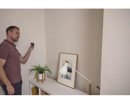 Ein Mann verwendet ein Ortungsgerät an einer Wohnzimmerwand neben einem Sideboard mit einer Pflanze und einer Tischleuchte.