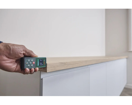 Eine Hand misst mit einem Bosch Laser Entfernungsmesser die Kante eines Holzelementes auf einem weißen Schrankmöbel.