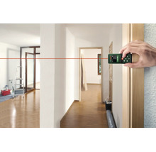 Eine Hand nutzt einen digitalen Bosch Laser Entfernungsmesser zur Messung in einem hellen Innenraum mit Holzboden.