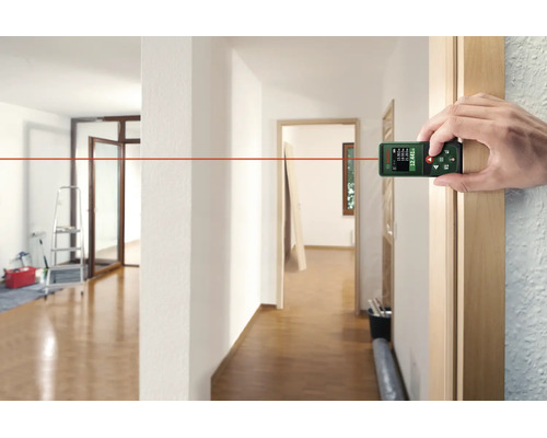 Eine Hand nutzt einen digitalen Bosch Laser Entfernungsmesser zur Messung in einem hellen Innenraum mit Holzboden.