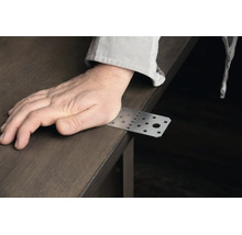 Eine Hand liegt auf einer Lochplatte aus Metall, die über die Kante einer Holzplatte ragt.