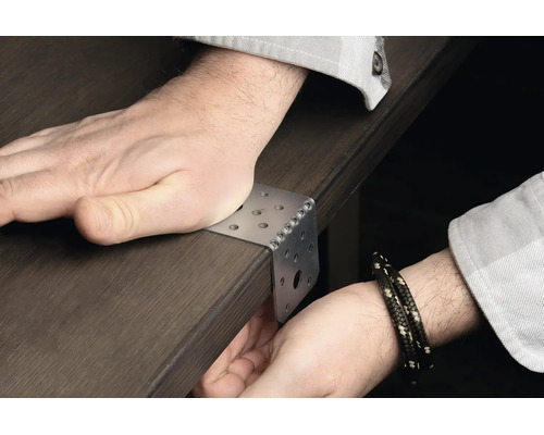 Ein Winkelverbinder aus Metall wird an einer dunklen Holzkante angesetzt und mit der Hand auf einer Tischplatte fixiert.