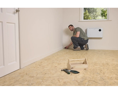 Ein Mann kniet auf einem OSB Platten Boden in einem Raum und montiert eine Fußleiste, neben ihm steht ein hölzerner Werkzeugkasten mit Werkzeug.