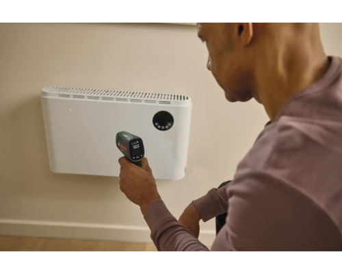 Eine Person misst die Temperatur eines Wandheizgerätes mit einem Bosch Infrarot Thermometer in einem Innenraum.