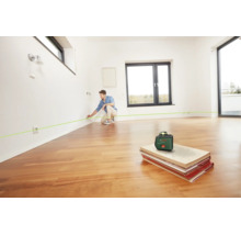 Ein Mann nutzt einen Bosch Kreuzlinienlaser in einem hellen, leeren Raum mit Parkettboden, um eine Linie an der Wand zu markieren.