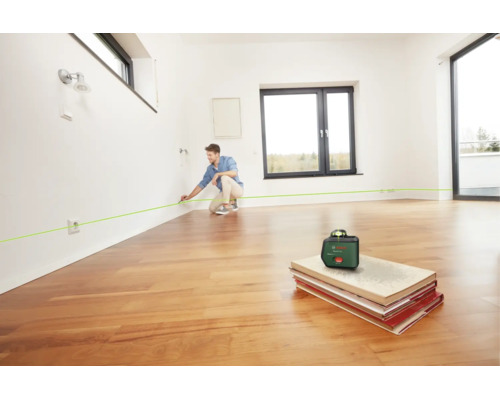 Ein Mann nutzt einen Bosch Kreuzlinienlaser in einem hellen, leeren Raum mit Parkettboden, um eine Linie an der Wand zu markieren.